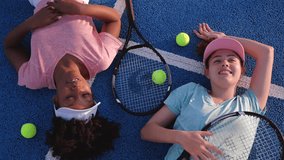 Two young women, tennis players, are lying on a blue tennis court, relaxing after a match, smiling and chatting, with their rackets and balls beside them - Powered by Shutterstock - Get 15% off with code: PIKWIZARD15
