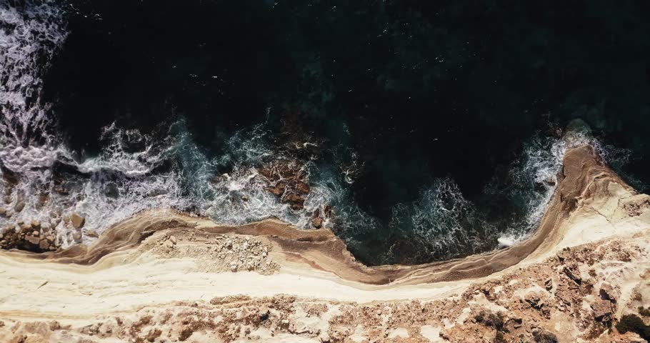 Drone top-down of waves crashing on rocky cliffs at sunset, Cyprus. Turquoise sea, rugged coastline, Mediterranean scenery, golden hour landscape.