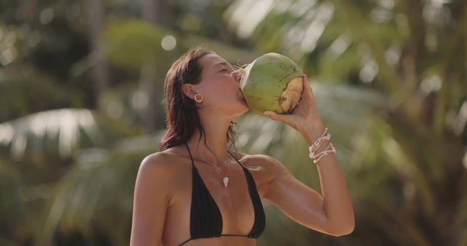 A young woman in a black bikini drinks fresh coconut water in a tropical setting, captured in slow motion. Perfect for travel, vacation, or tropical lifestyle promotions highlighting relaxation and
