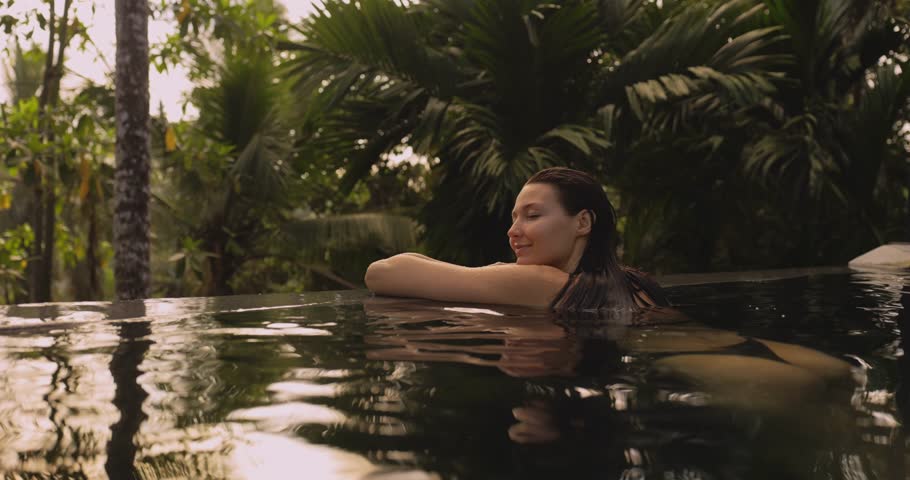 A serene woman in a bikini leans on the edge of an infinity pool, smiling as she enjoys the tranquil jungle surroundings. This scene embodies relaxation, luxury, and a peaceful tropical escape.