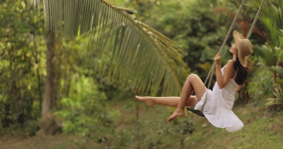 A carefree woman in a white summer dress and straw hat enjoys swinging beneath palm trees in a vibrant tropical garden. Perfect for concepts of relaxation, tropical travel, and outdoor adventure.