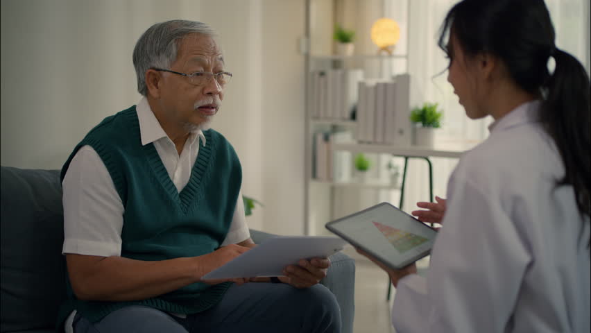 Elderly Asian man having health consultation with female doctor at home. Discussing nutrition chart using tablet and document.