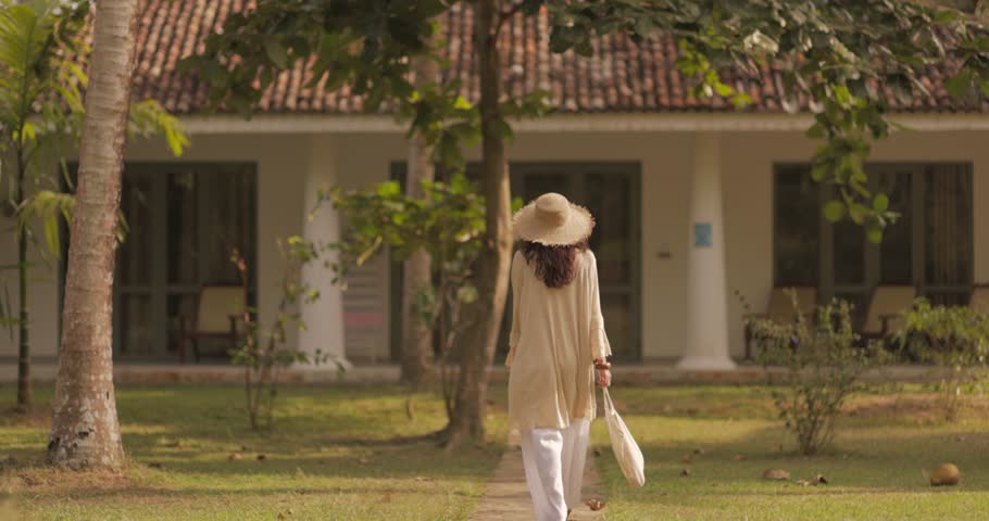 A woman in a chic, flowing outfit and straw hat walks slowly toward a tropical villa in slow motion. Ideal for concepts of expat living, luxury travel, and serene relaxation in tropical settings.