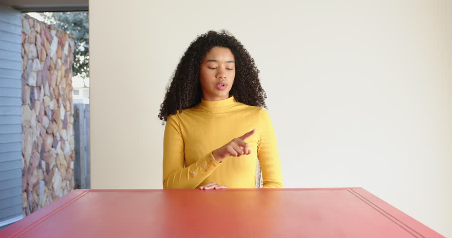 Close up of people talking to the camera, woman in yellow sweater sitting at red table. the person is fully visible, sitting, half body, pointing at things in the scene.