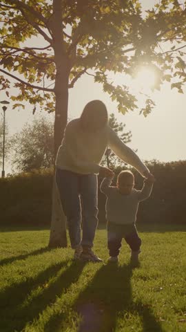 Vertical Screen: Woman helps her young boy as he takes his first steps on the lush grass of a park. The warm glow of the setting sun creates a heartwarming atmosphere.