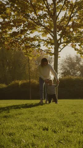 Vertical Screen: Mother watches with joy as her young child takes his first steps on a sunny day in the park. The child walks on green grass, reaching for her hand.