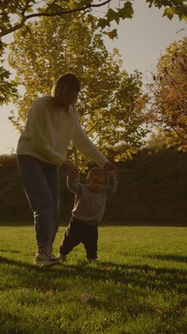 Vertical Screen: Boy enjoys learning to walk with the support of his mother in a lush green park during a bright afternoon. Sunlight filters through the trees as they share this special moment.