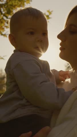Vertical Screen: Cheerful woman carries her child while walking through a park at sunset, embracing a moment of joy, love, and connection in the warm evening light.
