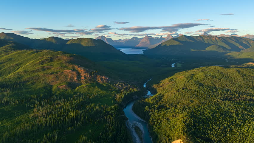 River Flowing Through Lush Green Hills Towards Mountain Lake At Sunset In Montana, USA. aerial shot, timelapse