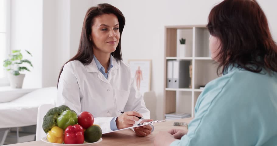 Pretty friendly nutritionist woman talking with her fat female patient and making notes in notepad about diet plan for weight loss sitting in office. Healthy food and nutrition concept. 4k video.