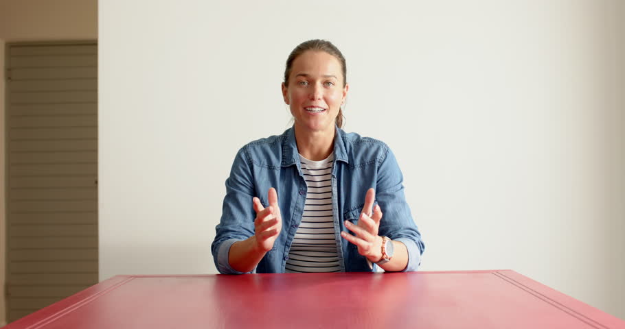 Close up of people talking to the camera, woman in denim jacket sitting at red table. the person is fully visible, sitting, half body, talking to the camera with exaggerated hand movements.