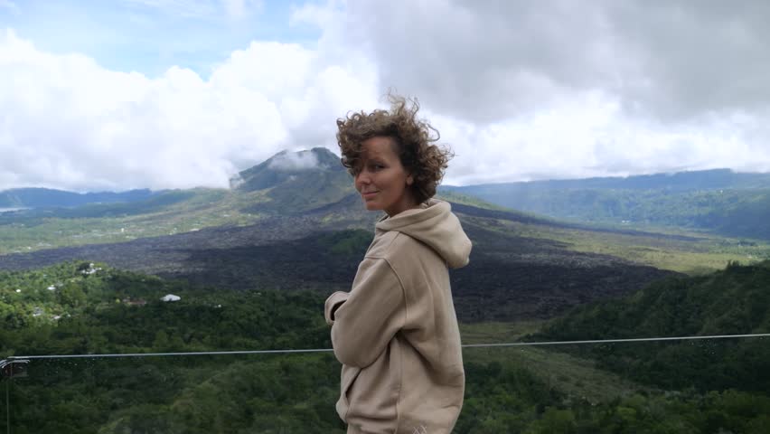 Young woman tourist with curly hair wearing beige hoodie stands with arms crossed, admiring the stunning volcanic landscape of mount batur in bali, indonesia