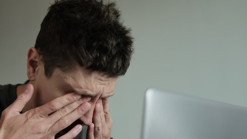 Man rubbing eyes while working on laptop. Tired eyes from long screen time. Freelancer blinking and massaging eyes. Vision problems from long hours on laptop