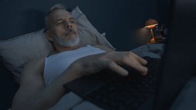 Rack focus shot of sleepless senior man lying in bed in dark room, surfing the Internet on laptop while suffering from insomnia during late night - Powered by Shutterstock - Get 15% off with code: PIKWIZARD15