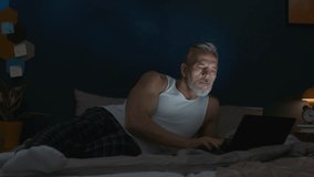 Senior Caucasian man in tank top and plaid pants lying on bed and browsing the internet on laptop in dimly lit bedroom at night - Powered by Shutterstock - Get 15% off with code: PIKWIZARD15