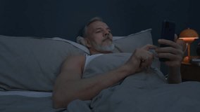 Sleepless senior man with grey hair and beard resting in bed and scrolling through social media on phone while suffering from insomnia at night - Powered by Shutterstock - Get 15% off with code: PIKWIZARD15