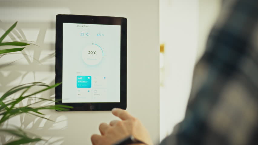 A person is using a touchscreen smart thermostat mounted on the wall in a living room. The individual is adjusting the temperature for optimal comfort. The setting is contemporary and bright