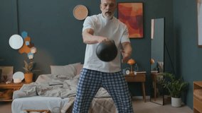 Strong senior man in pajamas performing kettlebell swing exercise in bedroom during home weightlifting workout - Powered by Shutterstock - Get 15% off with code: PIKWIZARD15