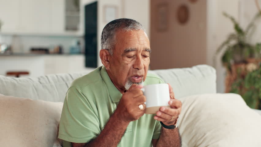Home, hot coffee and senior man on sofa, relax and blowing with espresso, morning and retirement. Pensioner, apartment and elderly person on couch, herbal tea and warm drink with wellness in lounge