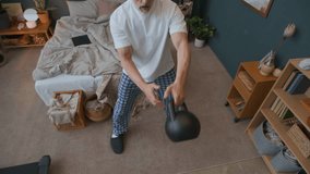 High angle view of senior man in pajamas and slippers standing in bedroom, doing kettlebell swings and one arm press while exercising at home - Powered by Shutterstock - Get 15% off with code: PIKWIZARD15