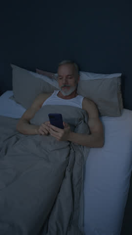 Vertical clip of senior Caucasian man lying under blanket in bed, scrolling on smartphone with smile in dim room during late night