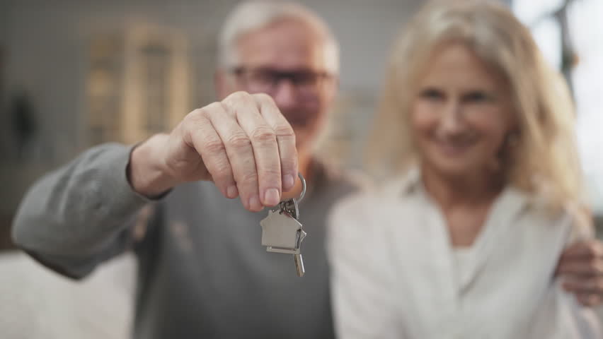 Buy new apartment, keys in hands of elderly man, blurred portrait of aged couple . Mature spouses rejoicing by finish of mortgage, happy house owners, real estate agency, rent, purchase or sale flat