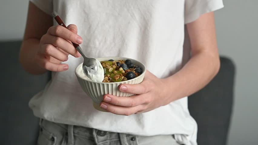 Bowl of granola with greek yogurt, kiwi and blueberries and spoon in hands of teenager in white t-shirt. Girl eating oat muesli. Healthy dairy breakfast concept.