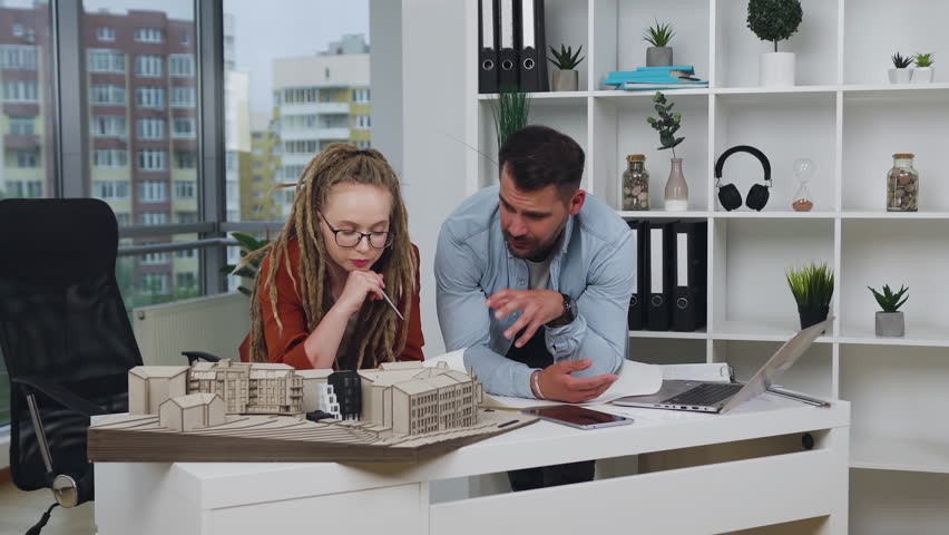 Man together with his stylish creative female colleague with dreadlocks discussing the model of buildings in architectural bureau