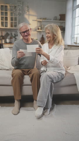 Aged people viewing old photographs, memories about happy life moments, vertical portrait in apartment. Happy aged spouses enjoying warm memories, smiling elderly people, calm and happy retirement