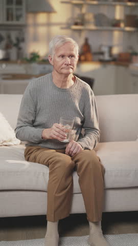 Old man drinking fresh water, healthy lifestyle and useful habits in old age. Prevention of dehydration, quench thirst, vertical video with single elderly man with glass of water sitting on couch
