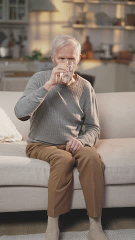Old man drinking fresh water, healthy lifestyle and useful habits in old age. Prevention of dehydration, quench thirst, vertical video with single elderly man with glass of water sitting on couch