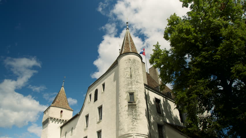 Wide shot of Château de Nyon from the west side with the flag waving, captured in a tracking and pan shot. A tree appears in the foreground, under a blue sky with clouds.