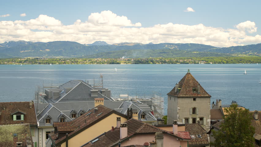 Wide, peaceful view of Lake Geneva with the town of Nyon on the shoreline. Boats, mountains in the distance, and partly cloudy skies are visible.
