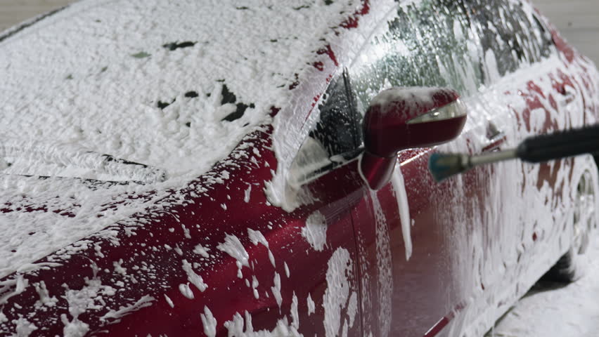 Side view of red car covered in soap foam being rinsed with high pressure water spray by someone in garage interior showing dynamic flow of water and dripping soap suds during professional cleaning
