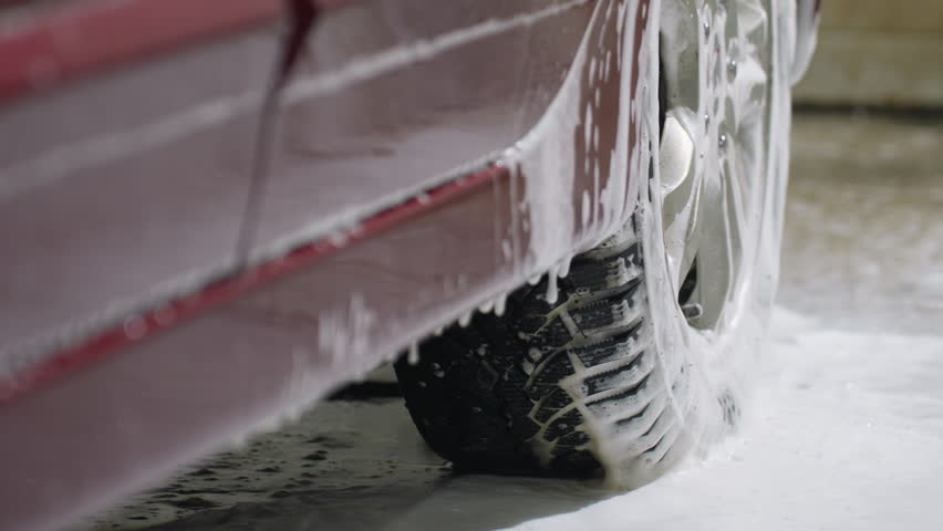 Close up of soapy water dripping from red car body near tire during washing process with suds and droplets creating dynamic texture and fluid flow on vehicle exterior inside garage environment