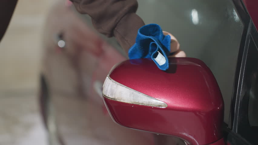 close up hand view of person wearing brown hoodie using blue cloth to clean shiny red car door near side mirror during detailing session inside garage under bright indoor lighting