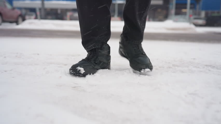 Close up lower angle view of person wearing black boots walking across snow covered ground with snowflakes falling and parked cars visible as he enter a car in background during cold winter day