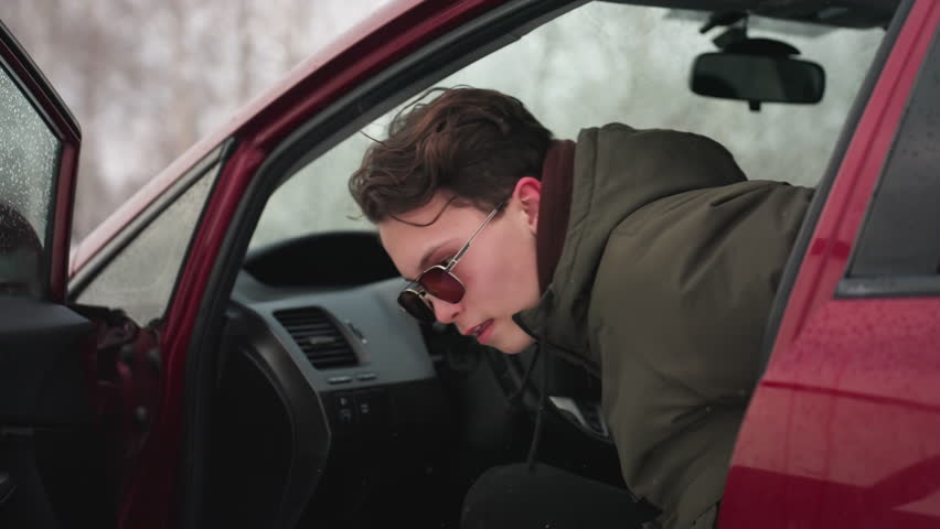 Man wearing winter jacket exits red car during cold weather with visible snowflakes in air and water droplets on vehicle, capturing moment of winter activity in outdoor snowy setting
