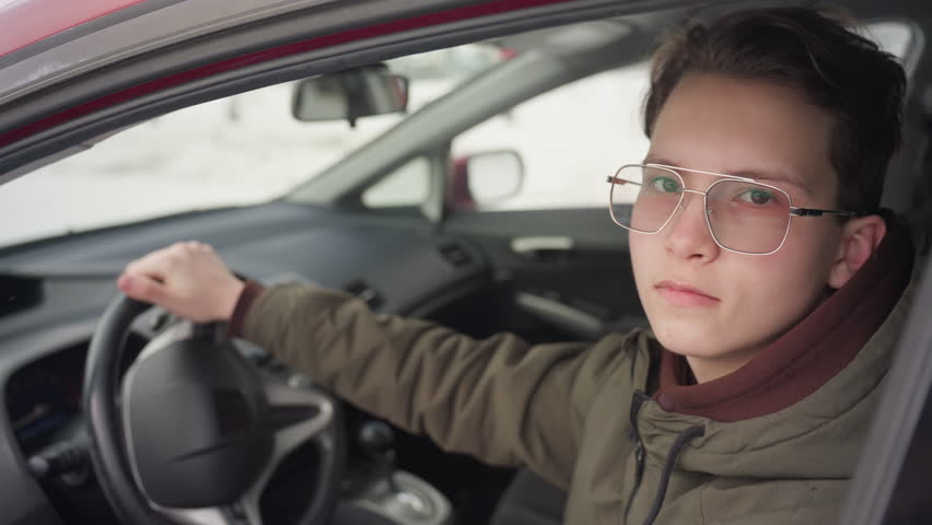boy in sunglasses inside car lowers driver window and gazes through raindrop-covered glass toward camera with cold snowy winter street visible in background during quiet moment