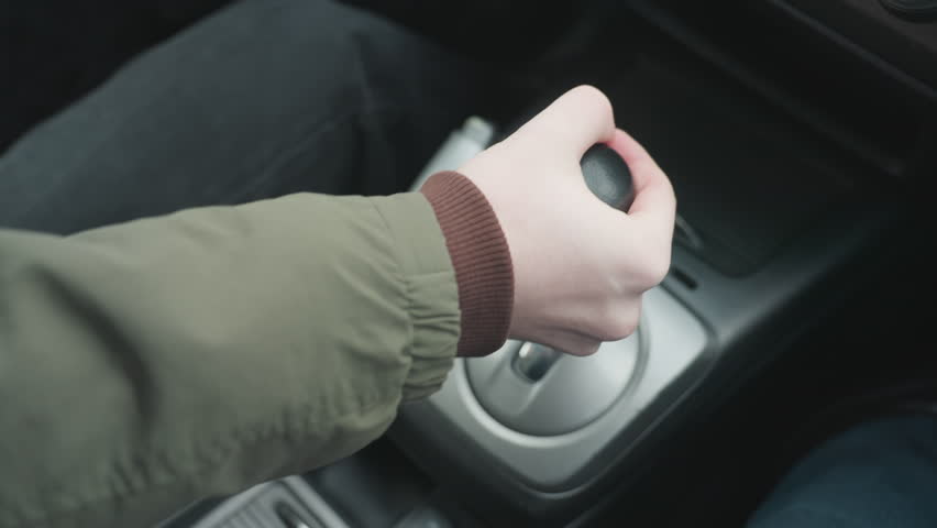 Close up top down view of driver pushing gear shift lever into park position in car center console showing hand grip on leather knob and metallic base under soft daylight in modern interior