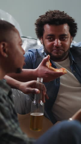 Young African-American builders eat pizza with beer and tell jokes sitting near couch in renovated room. Young black men celebrate repair finish in apartment
