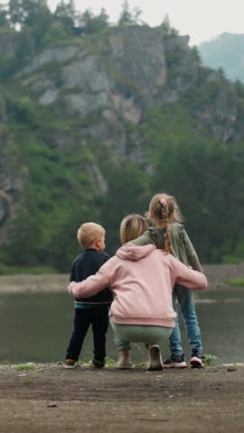 Mother hugs little children boy and girl spending time on bank of picturesque calm river at highland resort on cloudy summer day backside view