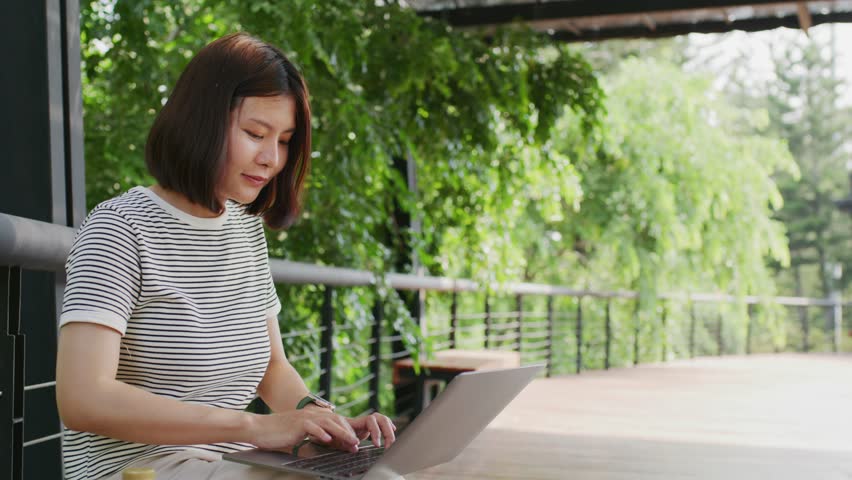 Young woman sitting on a bench with a laptop, working remotely in a green outdoor setting surrounded by trees and sunlight.