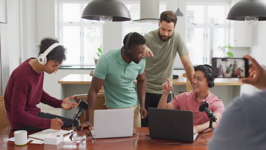 Five men recording podcast in kitchen using laptops and mics, showcasing floating emoji graphics. Collaboration, content creation, digital media, modern interior, casual workspace, technology,