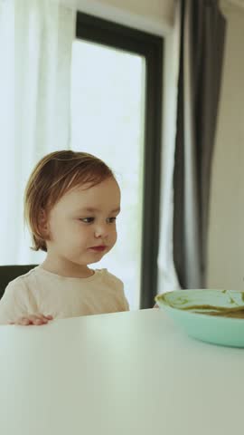  Vertical video: Child happily eats food from a spoon with his mouth wide open. A mother is feeding a girl sitting at a white table.