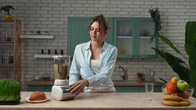 Home cooking and healthy eating concept. Young woman standing at the table in the kitchen preparing smoothie for breakfast, pushes button on blender and mixing fruits, positive expression. Slow motion - Powered by Shutterstock - Get 15% off with code: PIKWIZARD15