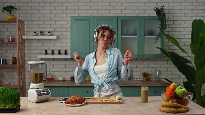 Home cooking and healthy eating concept. Young woman at the table in the kitchen preparing healthy breakfast, listens music in headphones on smartphone, dancing and drinking smoothie. Slow motion