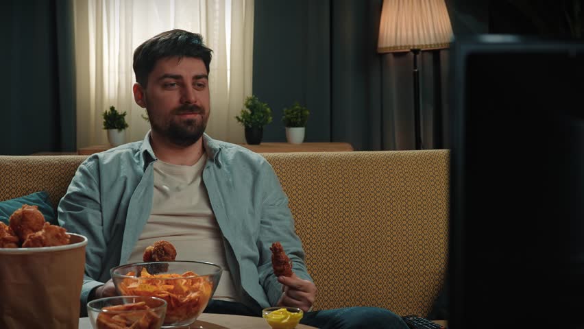 Junk food advertisement concept. Bearded man spending time at home with fast food delivery, sits on couch watching tv, holds friend chicken in hands smells and bites, enjoying the taste. Slow motion