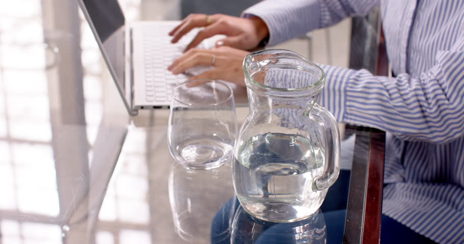 Smiling woman in office drinking water while working on laptop. Business, professional, hydration, technology, workplace, productivity