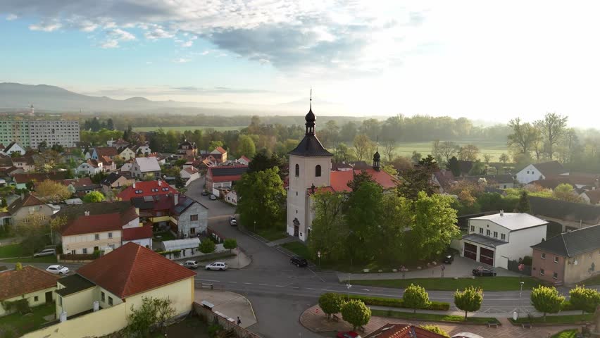 Drone footage captures a church (kostel) at sunrise, surrounded by soft morning fog. The golden light and mist create a magical, peaceful atmosphere over the quiet landscape.